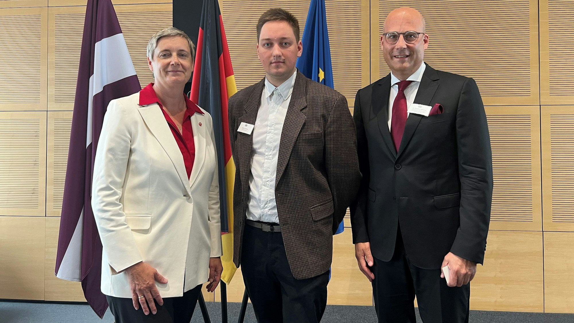 Eine Frau mit kurzen blonden Haaren und im weißen Blazer steht neben einem jungen Mann und einem mittelalten Mann im dunklen Anzug.