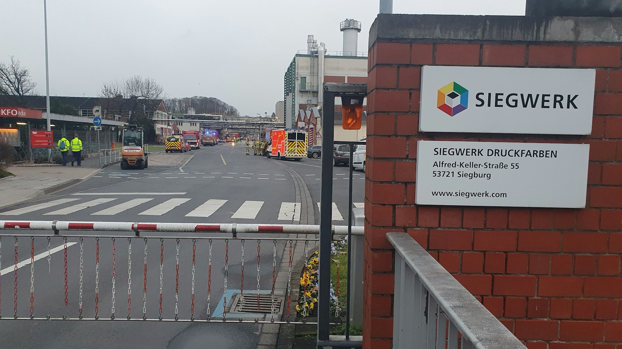 Feuerwehrleute und Einsatzfahrzeuge stehen auf dem Betriebsgelände hinter einer Schranke.