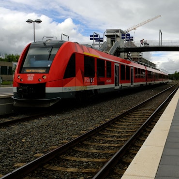 Die Fußgängerbrücke am Bahnhof Mechernich ist zu sehen. Ein roter Zug steht auf den Gleisen.
