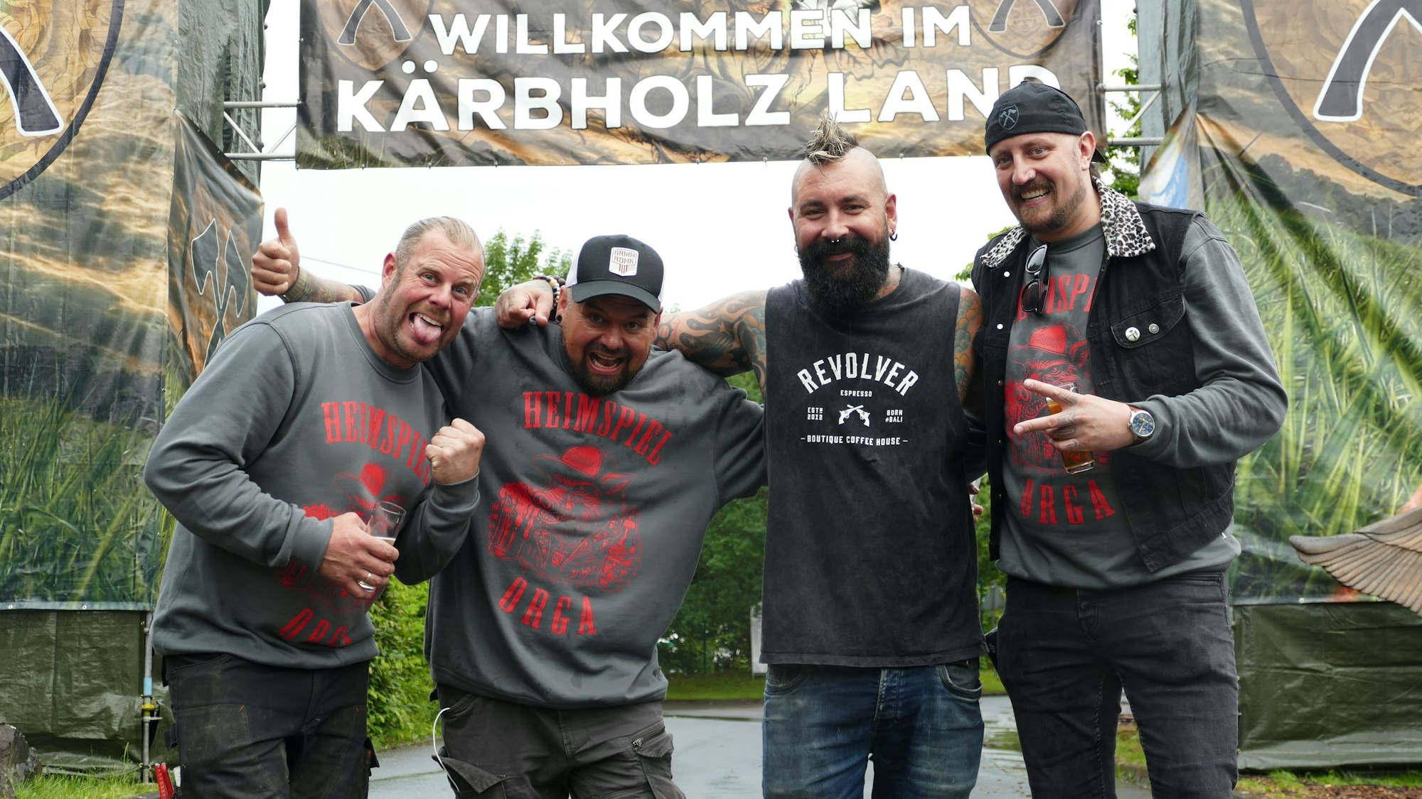 Vier Männer stehen unter einem Banner, auf dem steht: „Willkommen im Kärbholz-Land“.