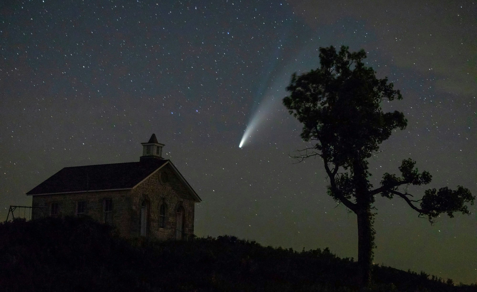 Komet Neowise oder C/2020 F3 ist am Nachthimmel über dem US-Reservat «Tallgrass National Prairie Preserve» zu sehen. Im Vordergrund des Fotos steht links ein Haus und rechts ein Baum.