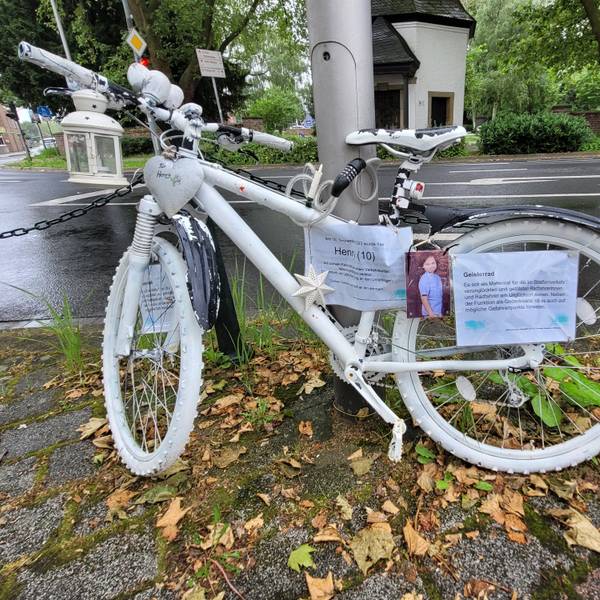 Ein weißes Fahrrad, Geisterfahrrad genannt, lehnt an einer Straßenlaterne.