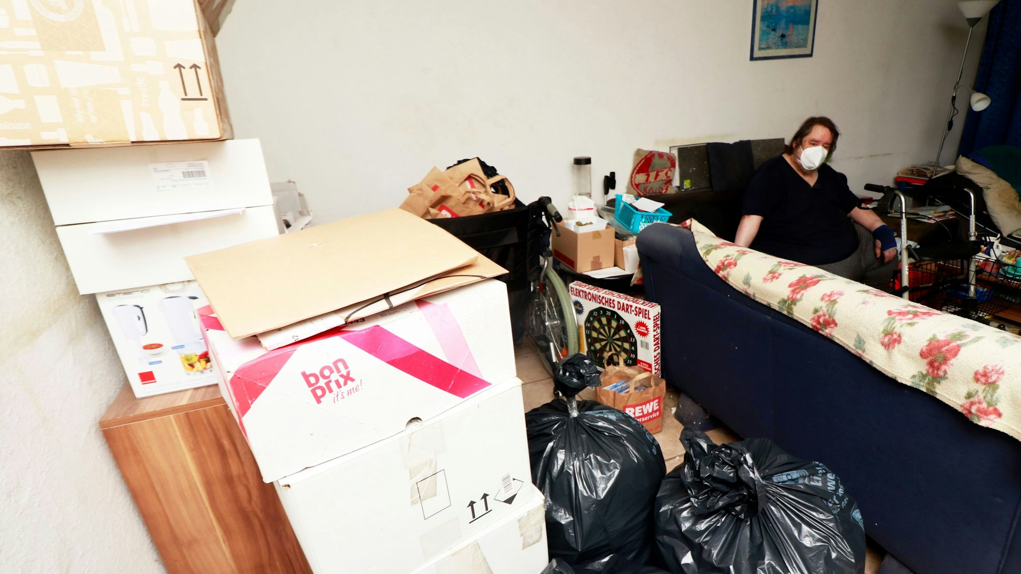 Köln: Müllsäcke und Packete prägen das Bild in dieser Wohnung. Ein Pflegebedürftiges Paar lebt in unwürdigen Zuständen und bekommt keine Hilfe von der Stadt und Pflegedienste. Zu sehen sind Müllsäcke und Umzugskartons, ein Mann mit Maske sitzt auf dem Sofa und stützt sich auf einen Rollator.