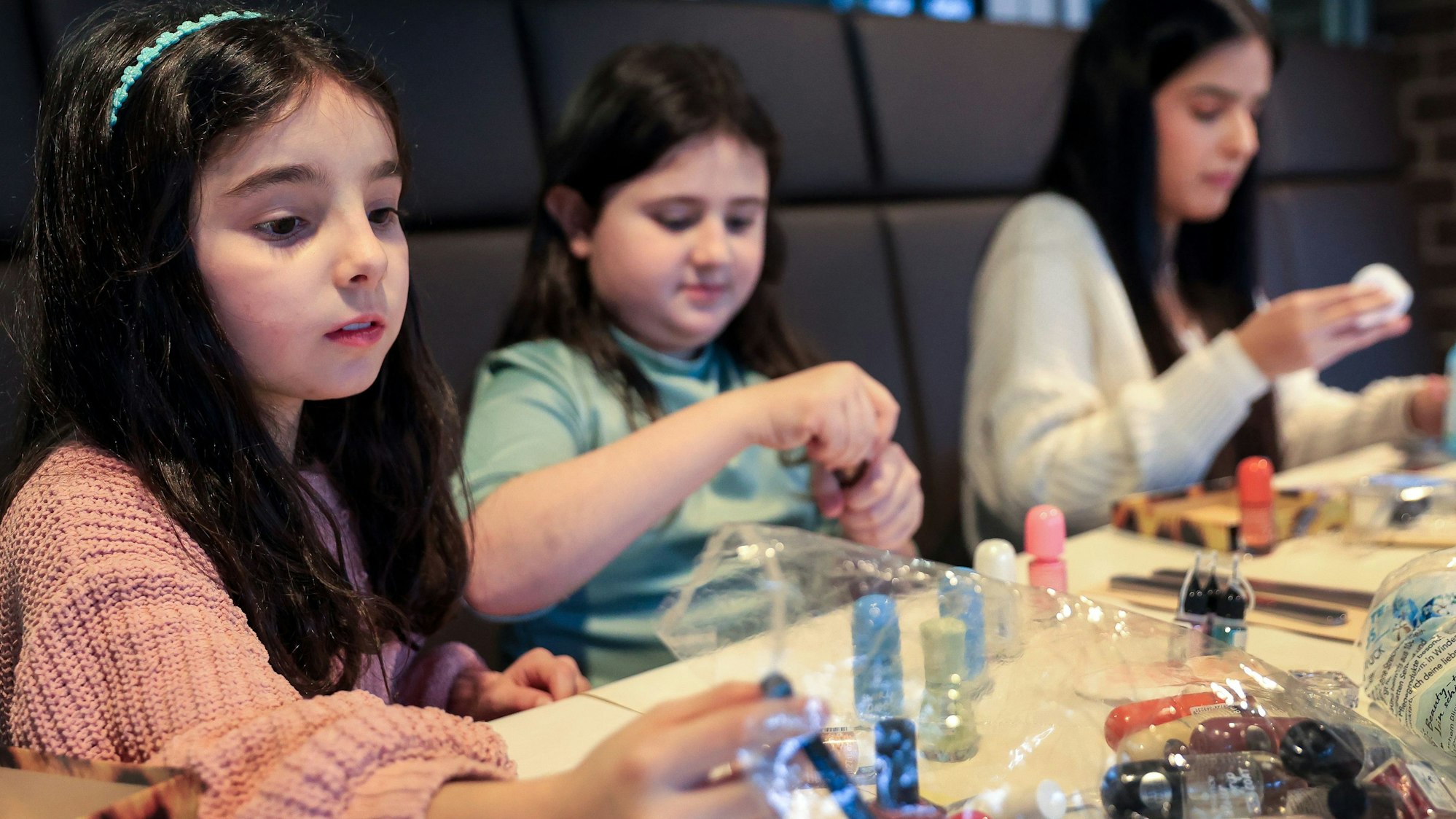 Drei junge Mädchen mit langen, braunen Haaren sitzen an einem großen Tisch im Bistro der Glashütte Porz und lackieren sich die Fingernägel