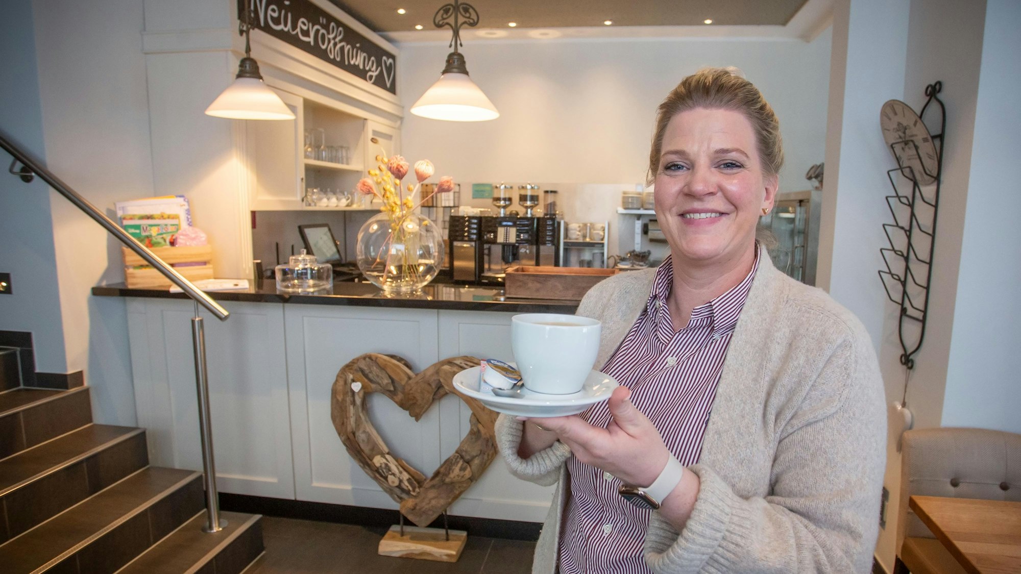 Eine Frau schaut in die Kamera, sie hat eine Tasse Kaffee mit Untertasse i der Hand, im Hintergrund sind ein Tresen und eine Kaffeemaschine zu sehen. Auf einer Tafel im oberen Bildrand steht das Wort „Neueröffnung“.