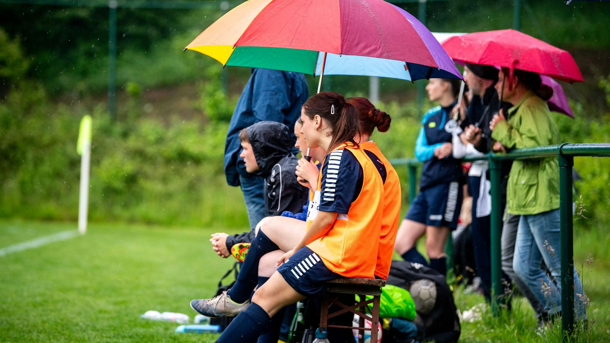 Das Bild zeigt die Auswechselbank des VfL Kommern. Einige Spielerinnen sitzen unter einem bunten Regenschirm.
