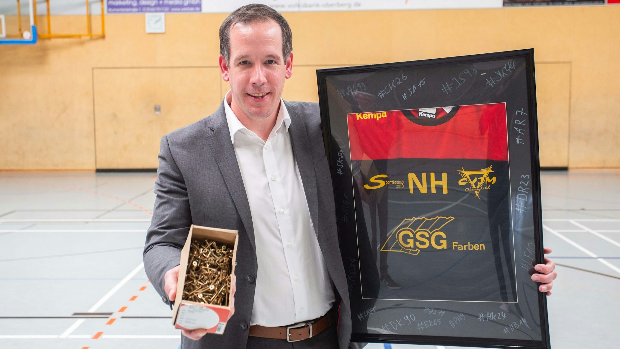 Nils Hühn hält ein eingerahmtes Handballtrikot und eine Koste voller Schrauben in den Händen. Er steht in einer Sporthalle.