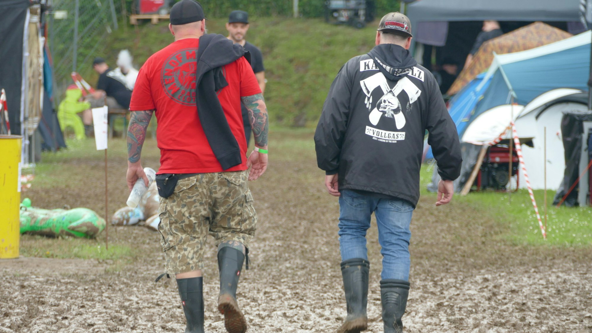 Zwei tätowierte Männer in Gummistiefeln waten durch den Schlamm auf einem Campingplatz mit Zelten. Einer trägt eine Kärbholz-Jacke.