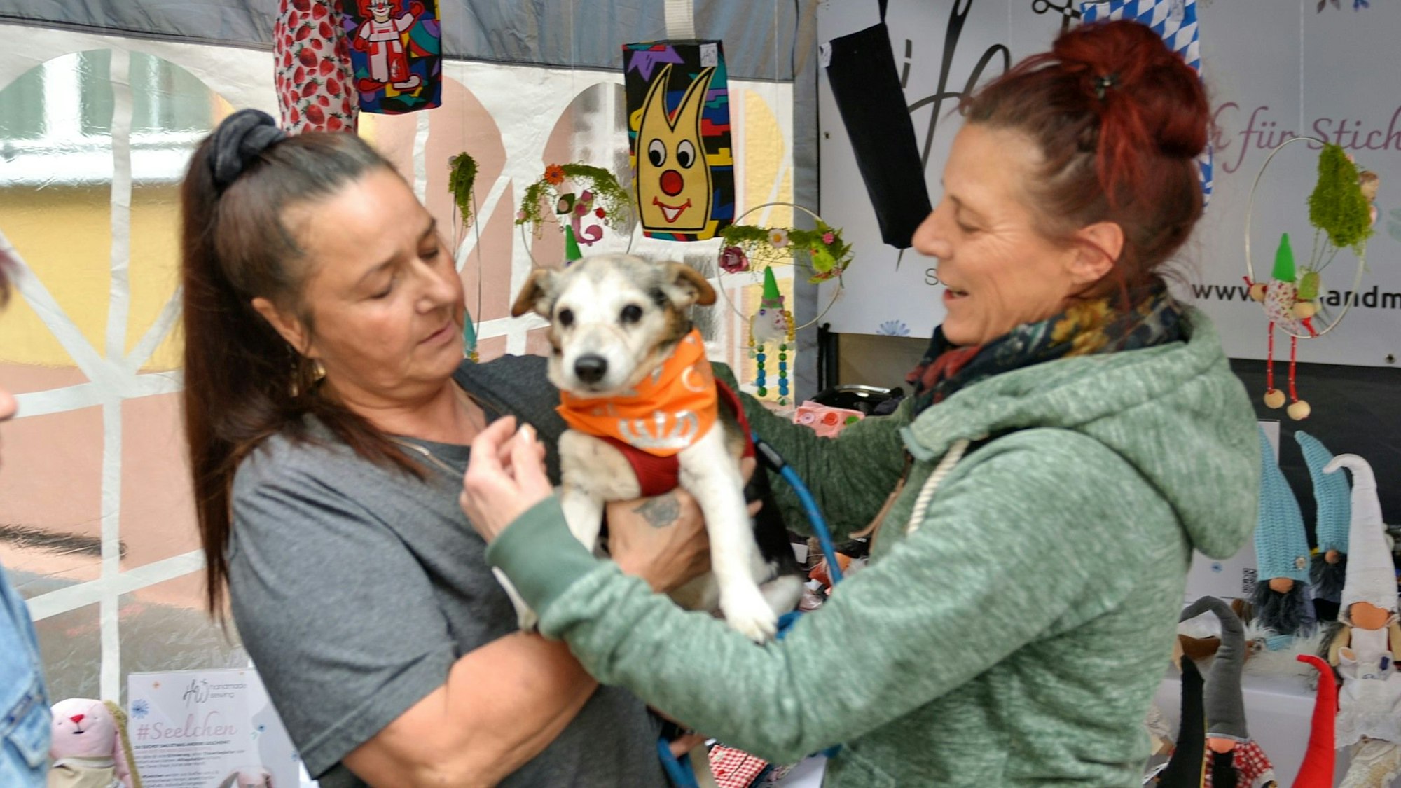 Eine Frau hält ihren kleinen Hund auf dem Arm, eine andere legt ihm ein Halstuch an.