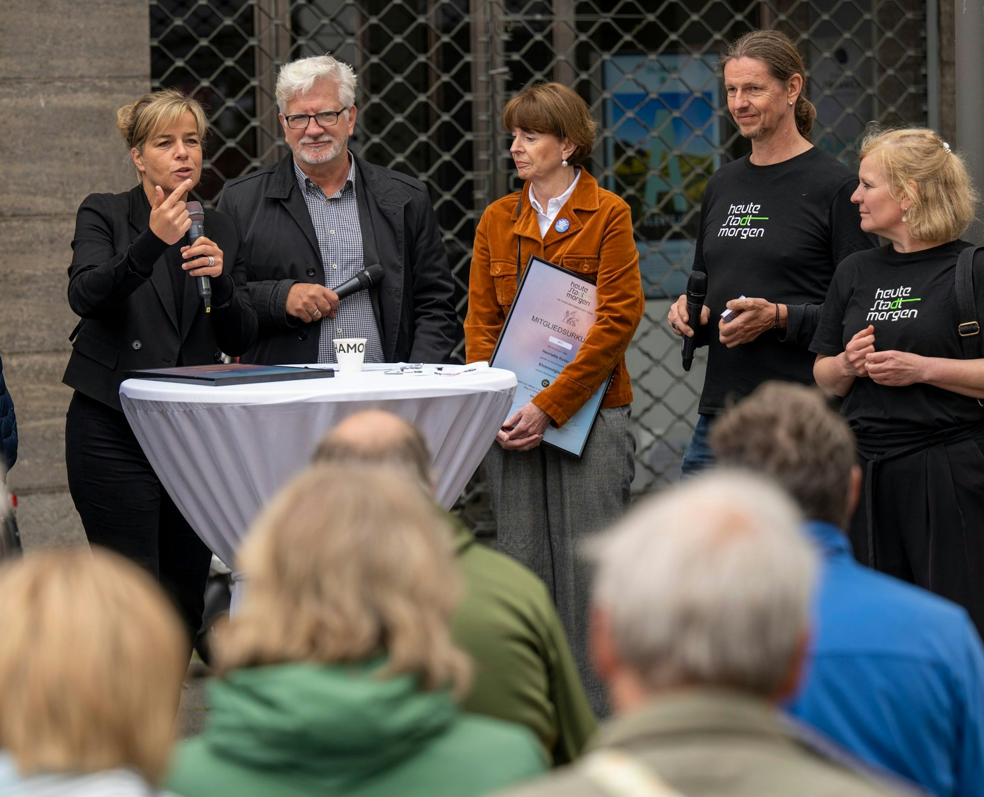 Menschen stehen an einem Stehtisch, eine Frau spricht in ein Mikrofon. Im Vordergrund sind sitzende Menschen von hinten zu sehen.