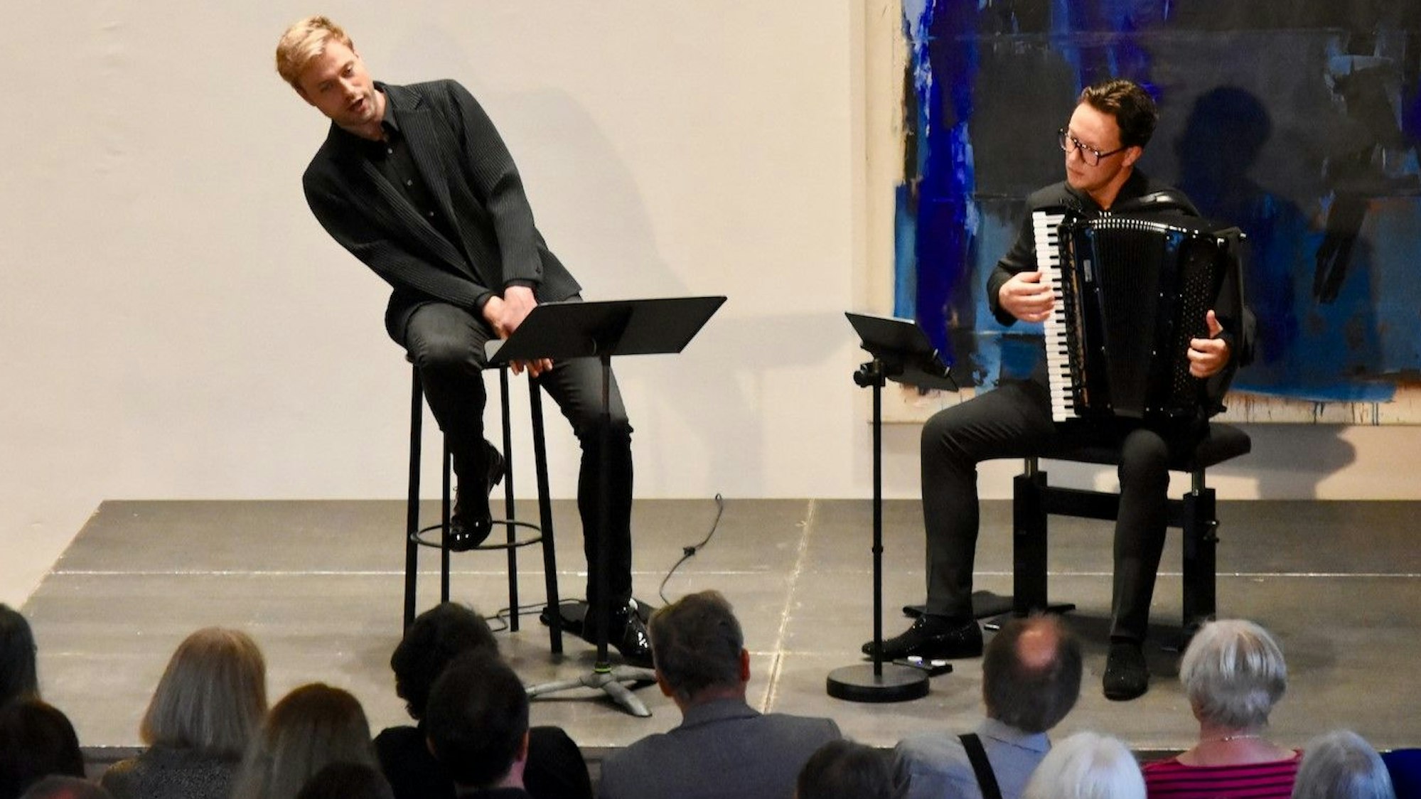 Ein blonder Sänger auf einem Barhocker und ein Mann mit Akkordeon auf einer Konzertbühne.