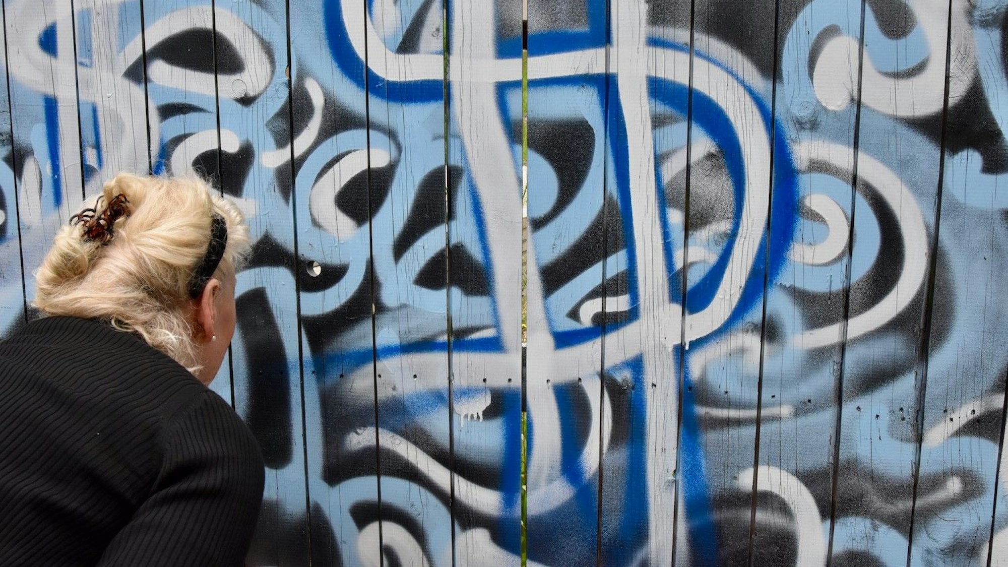 Eine blonde Frau schaut durch ein Loch in einem bunt bemalten Bauzaun.