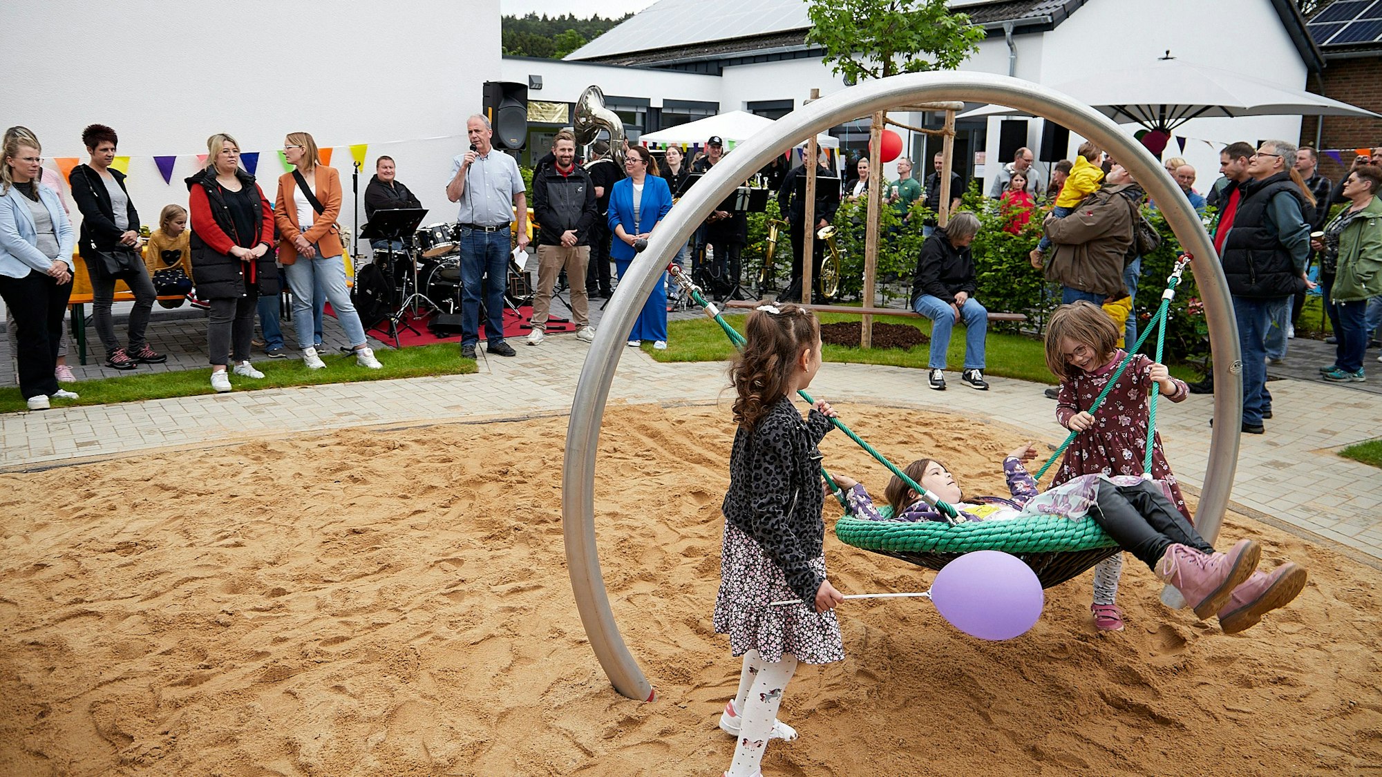 Drei Mädchen probieren die neuen Spielgeräte auf dem Spielplatz am Kindergarten in Sötenich aus. Im Hintergrund stehen zahlreiche Menschen.