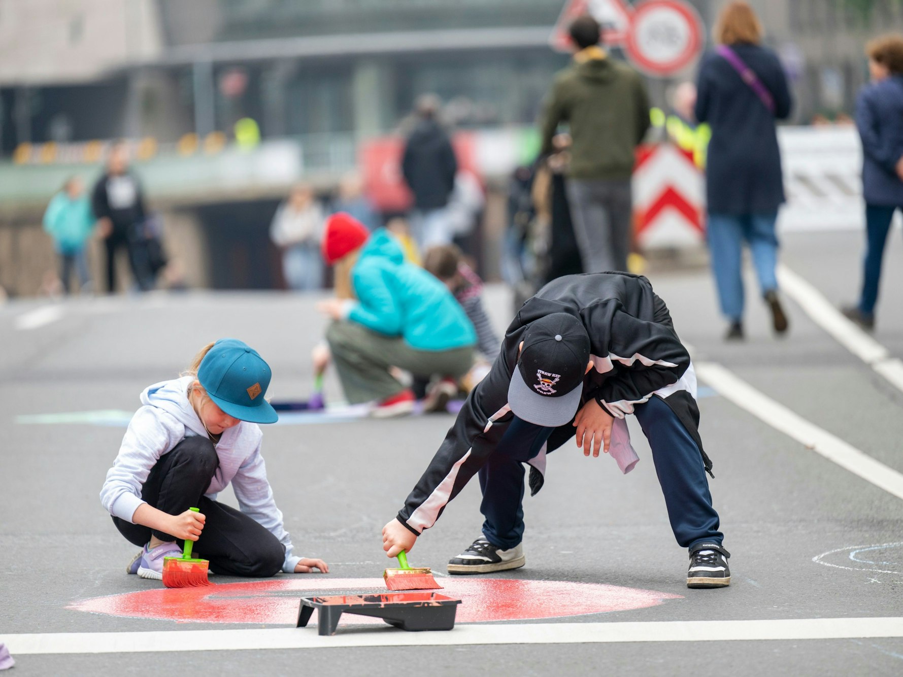 Kinder malen bunte Farben auf eine Straße. Im Hintergrund sind weitere Menschen zu sehen.