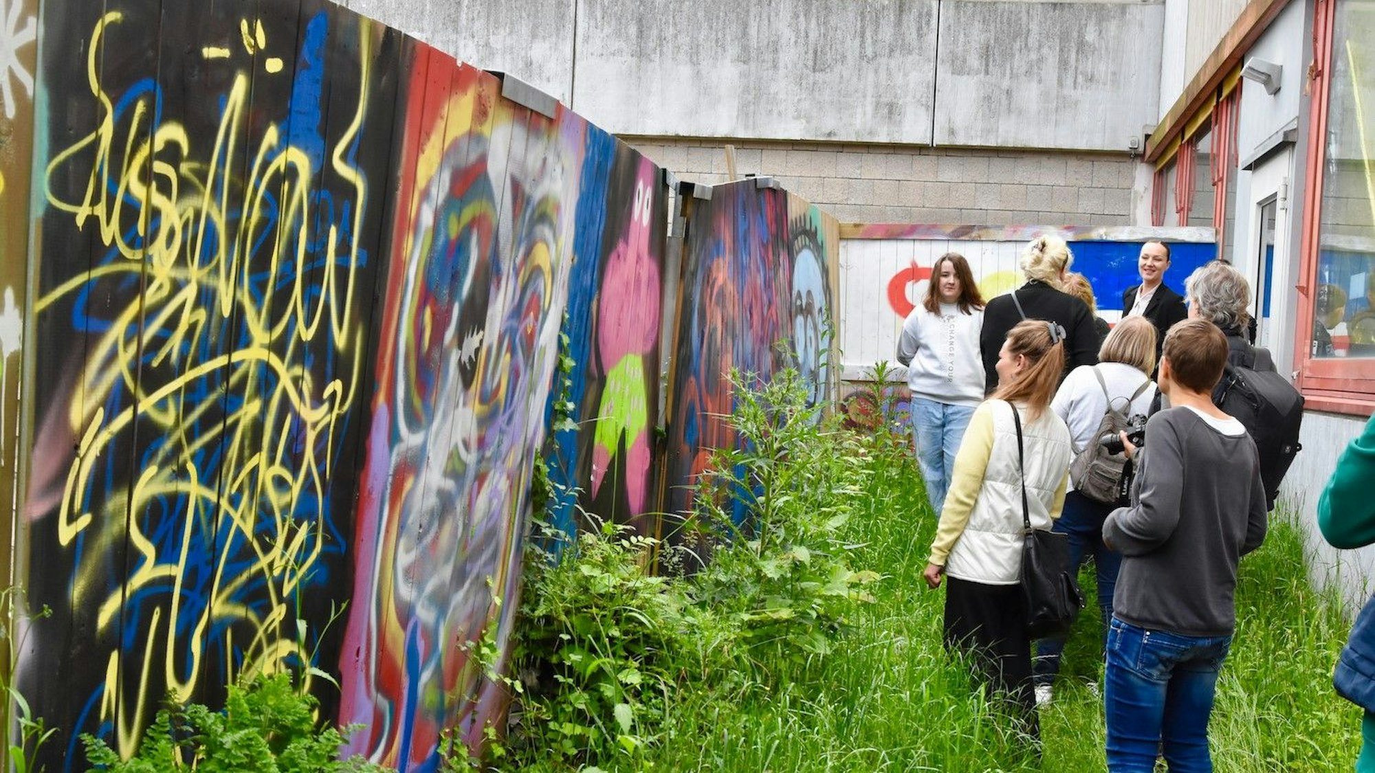 Eine Gruppe von Menschen betrachtet Graffiti-Kunst auf einem Bauzaun. Sie stehen in hohem Gras.