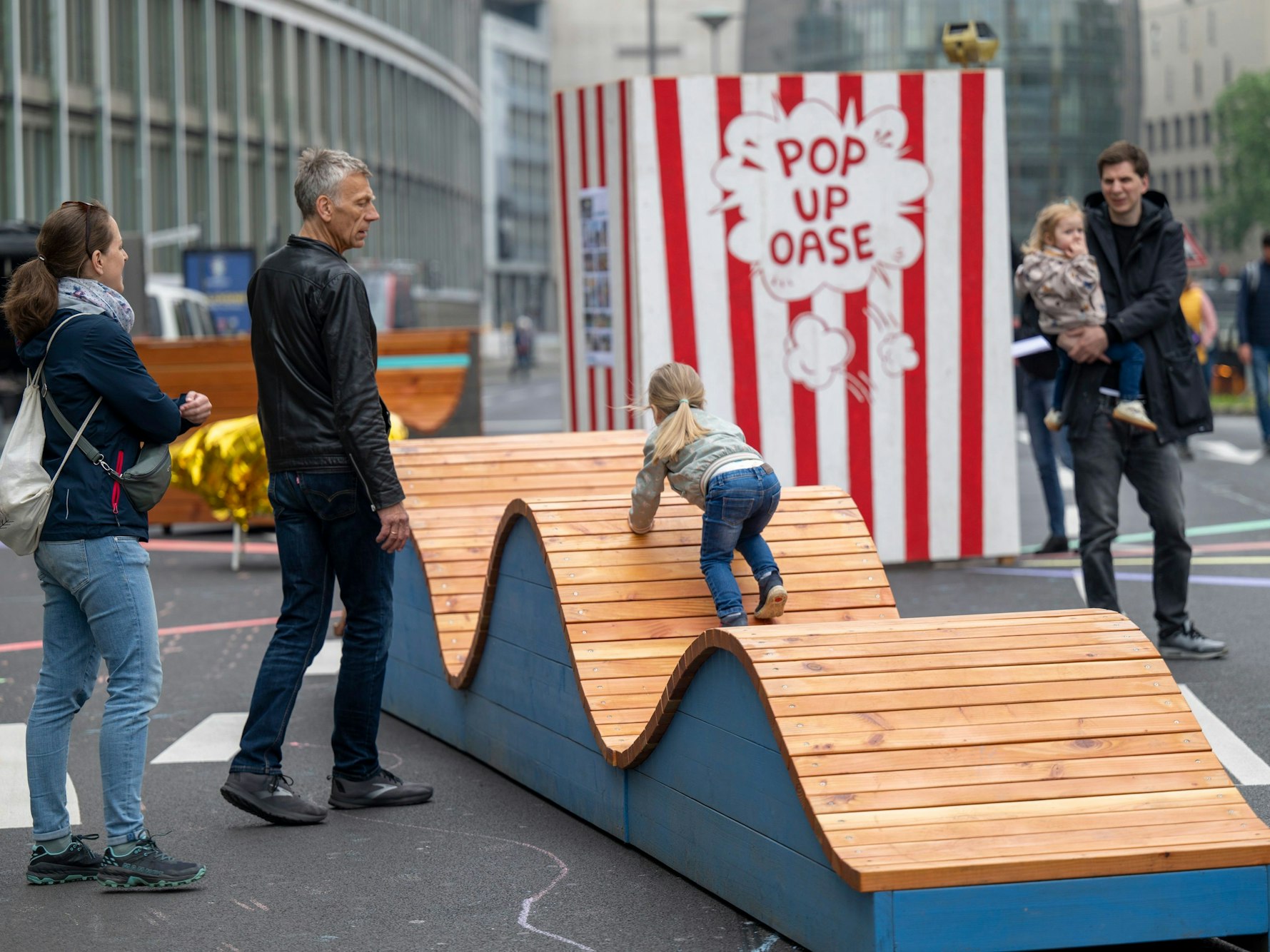 Ein Kind klettert auf einem hölzernen Sitzmöbelstück auf einer Straße. Daneben stehen meherere Menschen.