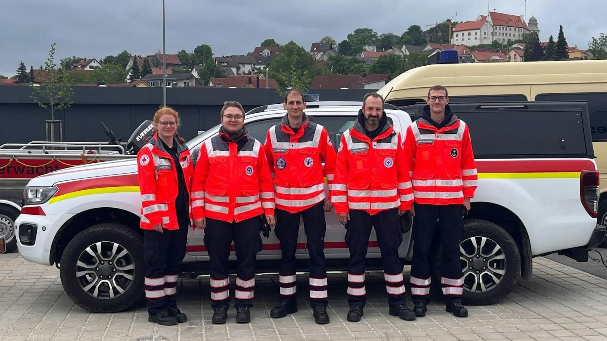 Fünf der insgesamt sieben Wasserwachtler des DRK, die aktuell in Landau an der Isar im Einsatz sind, stehen in ihrer Einsatzkleidung mit dunklen Hosen und roten Jacken vor einem Fahrzeug.