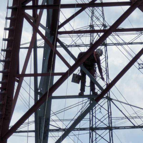 Ein Arbeiter mit Farbeimer und Pinseln streicht eine Strebe hoch oben in einem Stahlgittermast.