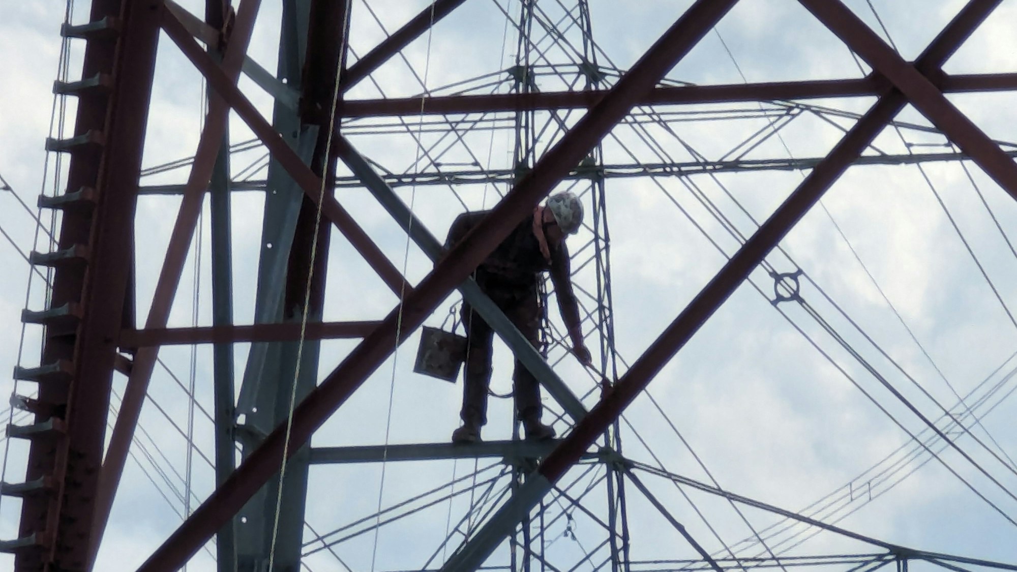 Ein Arbeiter mit Farbeimer und Pinseln streicht eine Strebe hoch oben in einem Stahlgittermast.