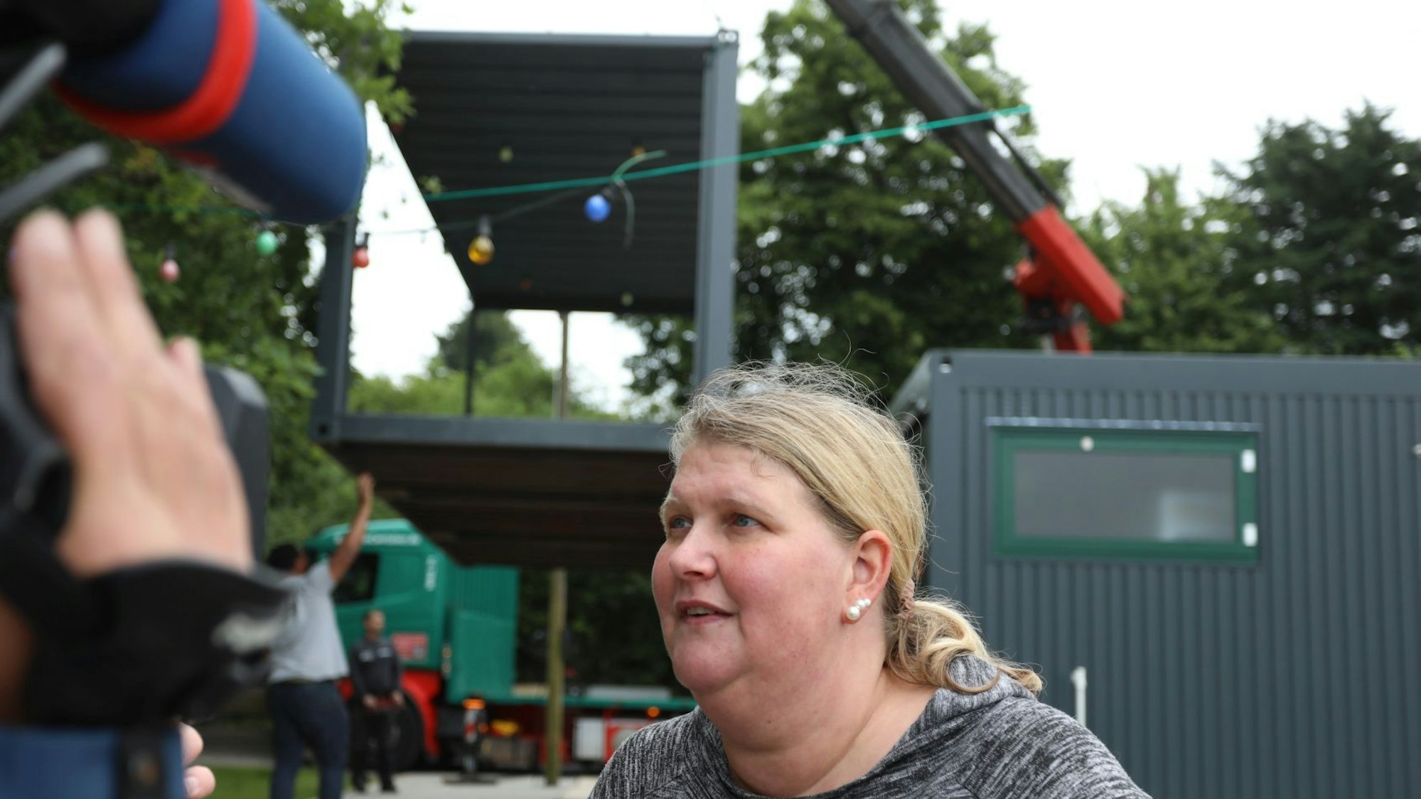 Eine Frau spricht in eine Fernsehkamera, im Hintergrund schwebt ein Container an einem Kran.