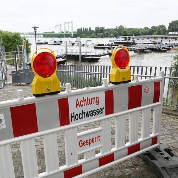 Ein Weg ist mit dem Schild „Achtung Hochwasser“ abgesperrt. Im Hintergrund ist der Rhein zu sehen.