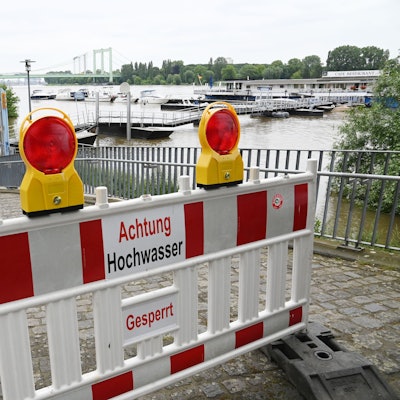 Ein Weg ist mit dem Schild „Achtung Hochwasser“ abgesperrt. Im Hintergrund ist der Rhein zu sehen.