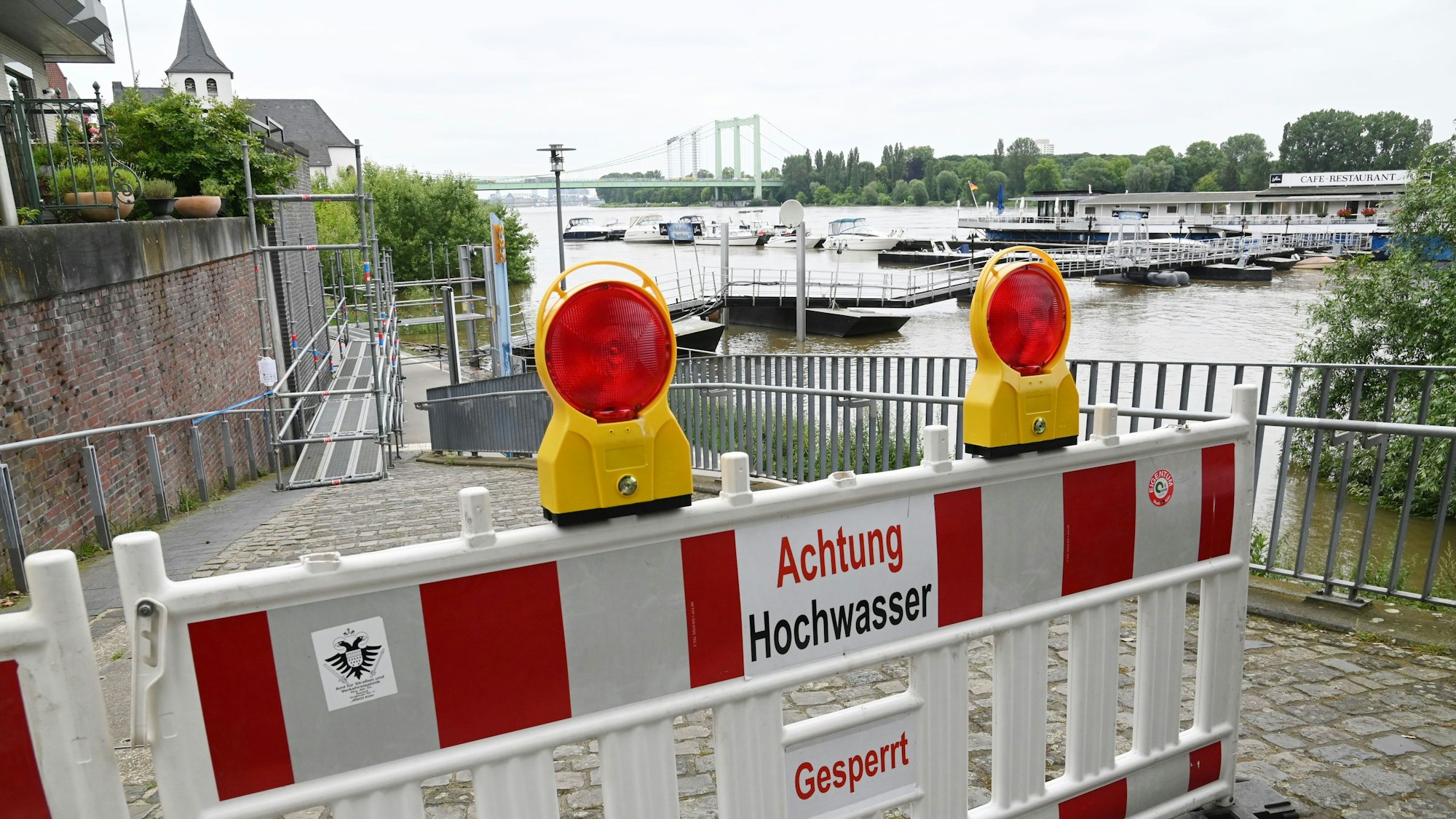 Ein Weg ist mit dem Schild „Achtung Hochwasser“ abgesperrt. Im Hintergrund ist der Rhein zu sehen.