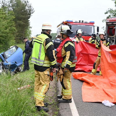 Ein blaues Auto liegt links im Straßengraben, der Fahrer ist bei dem Unfall ums Leben gekommen. Rechts sind Feuerwehrleute zu sehen, die rote Planen als Sichtschutz hochhalten.