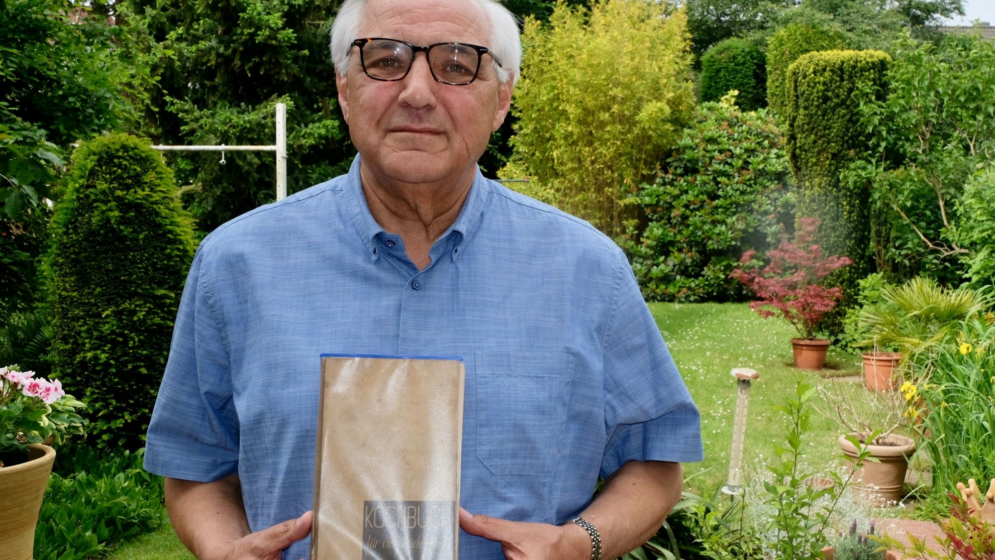 Zu sehen ist ein Mann, der ein Kochbuch hält, in einem Garten.