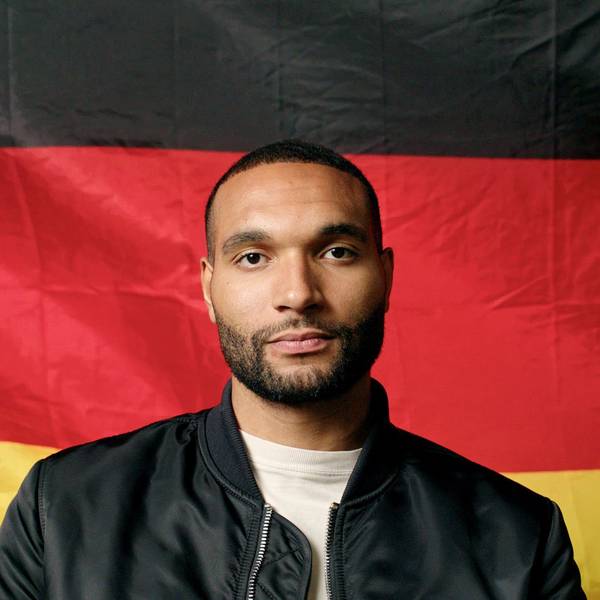 Nationalspieler Jonathan Tah sitzt vor einer Deutschlandflagge. Er trägt ein weißes T-Shirt und eine schwarze Jacke und blickt in die Kamera.
