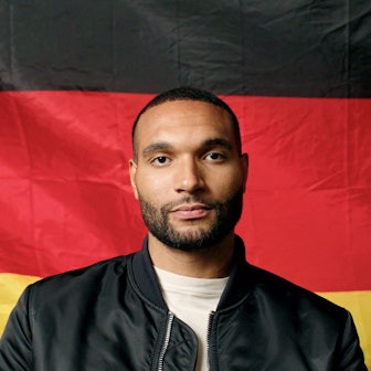 Nationalspieler Jonathan Tah sitzt vor einer Deutschlandflagge. Er trägt ein weißes T-Shirt und eine schwarze Jacke und blickt in die Kamera.