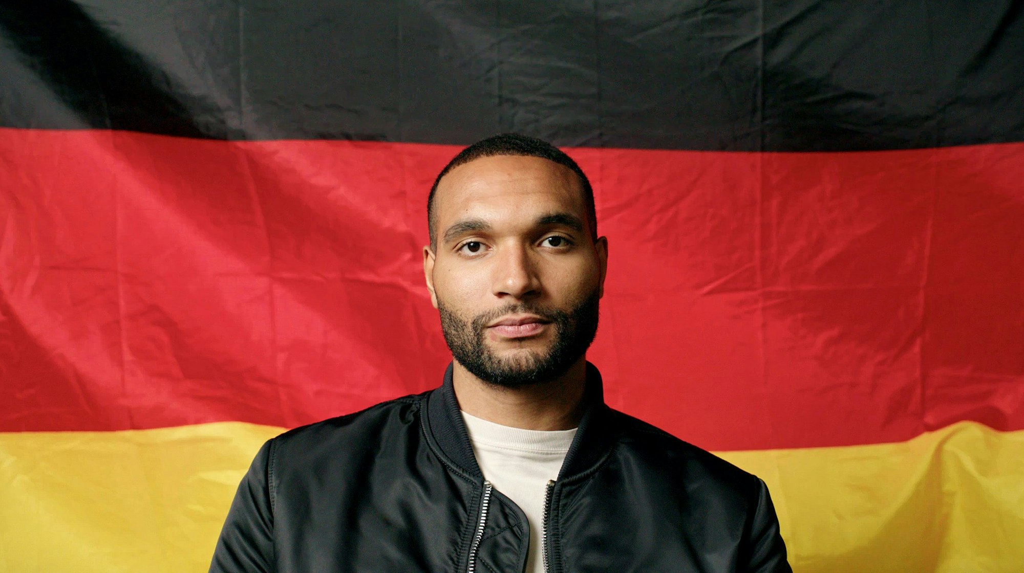 Nationalspieler Jonathan Tah sitzt vor einer Deutschlandflagge. Er trägt ein weißes T-Shirt und eine schwarze Jacke und blickt in die Kamera.