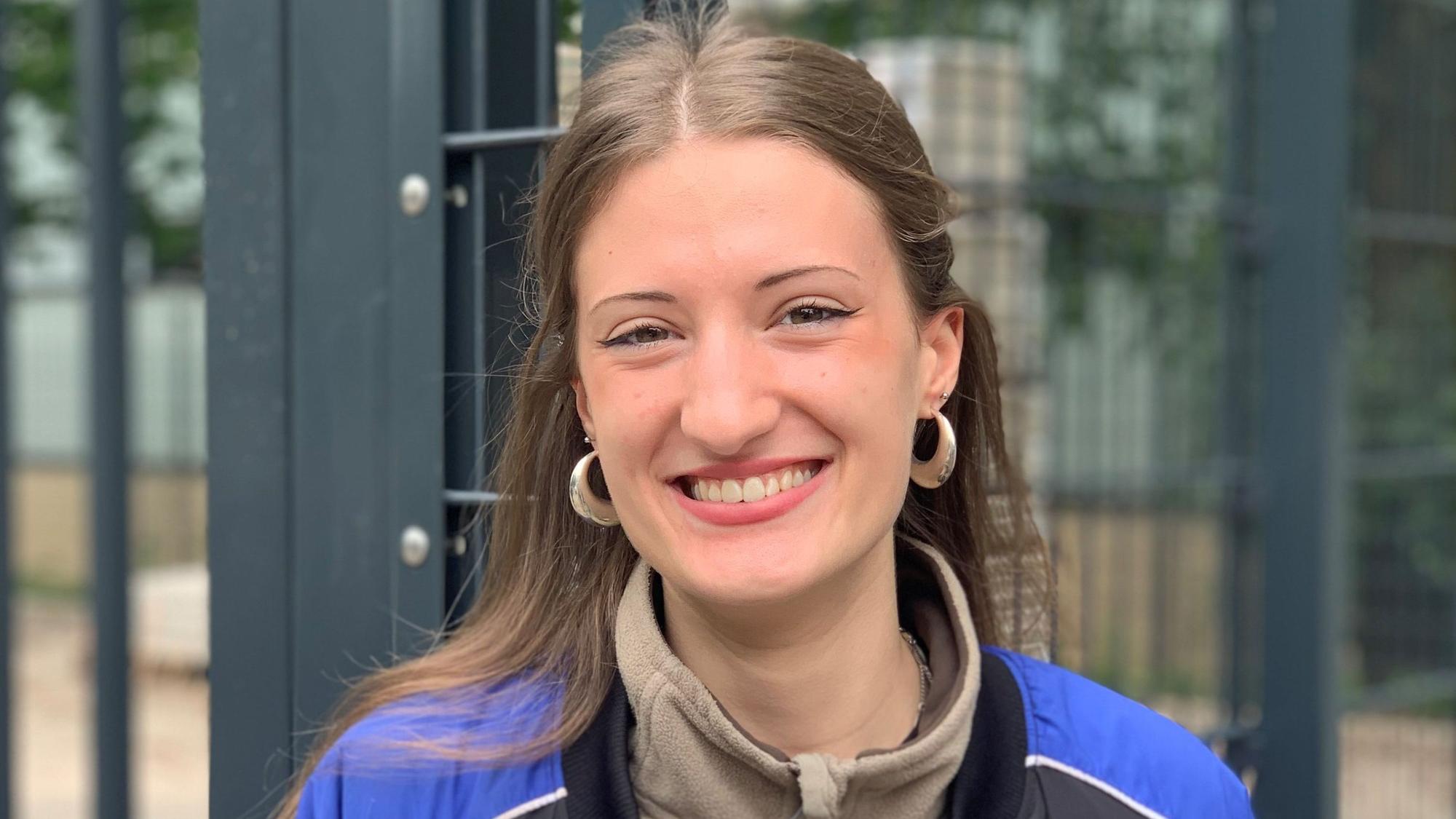Eine junge Frau steht vor einem Schulgebäude und lächelt.