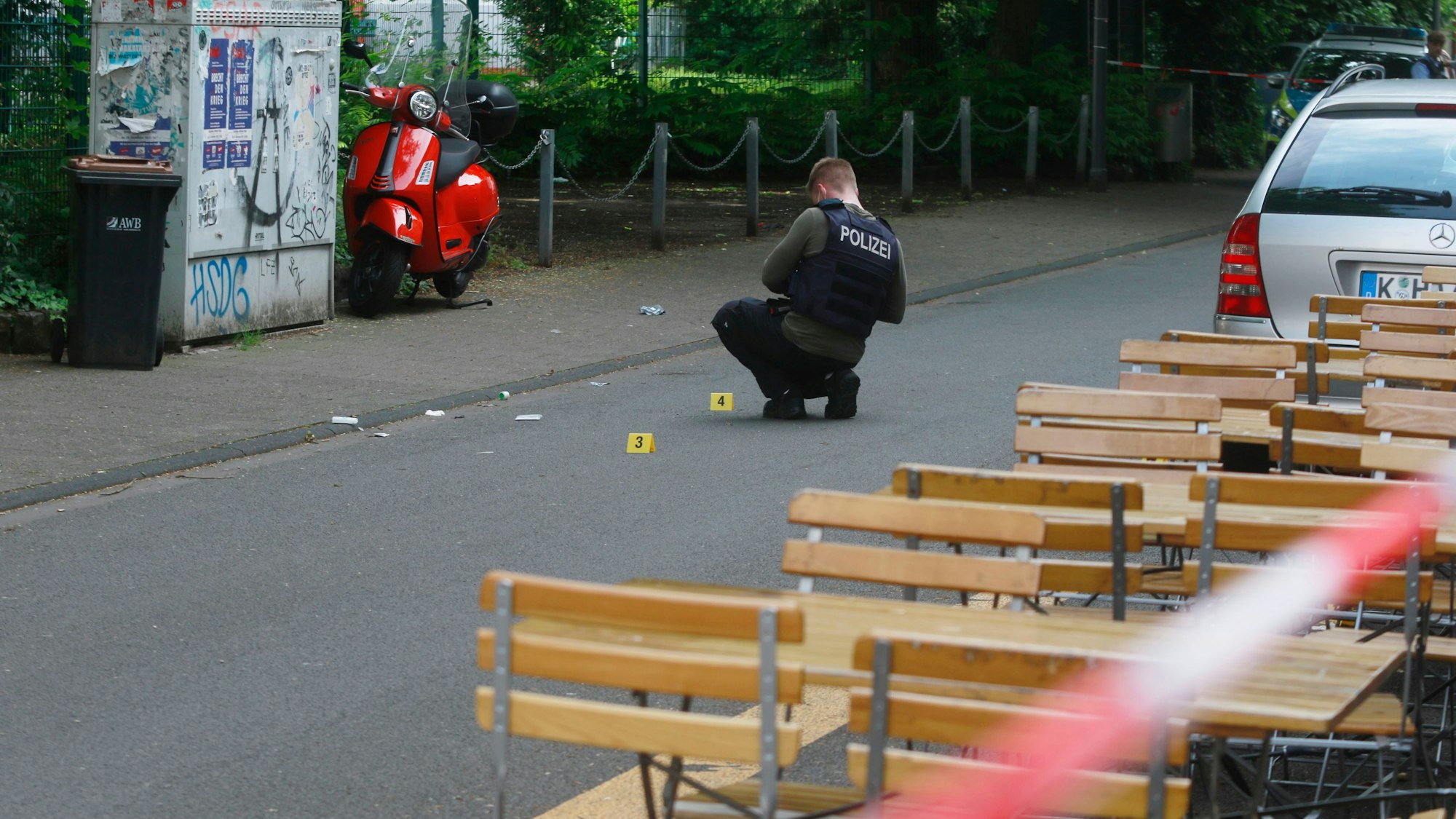 Ein Polizist untersucht einen Tatort auf der Straße, umgeben von einem Absperrband, einem Auto und Tischen.