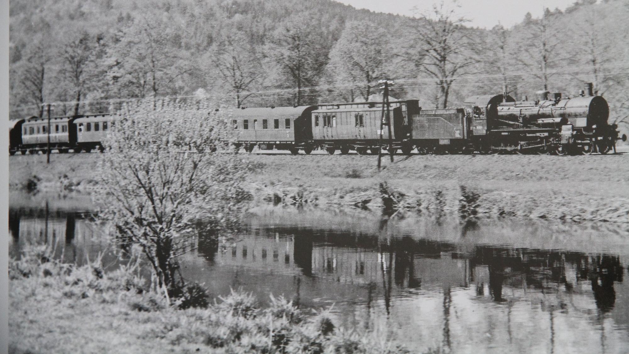 Eine Dampflok mit Personenwagen fährt eine Eisenbahnstrecke an einem Flussufer entlang