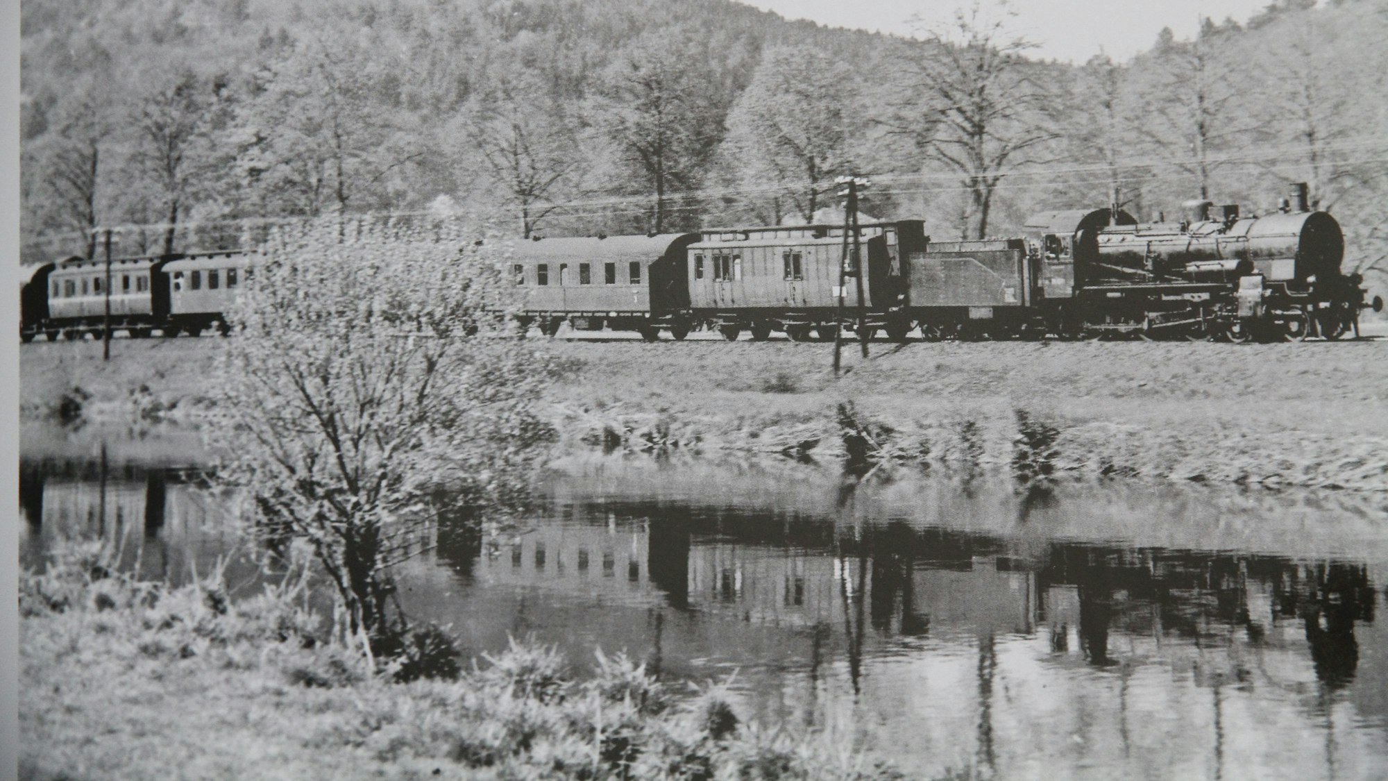Eine Dampflok mit Personenwagen fährt eine Eisenbahnstrecke an einem Flussufer entlang
