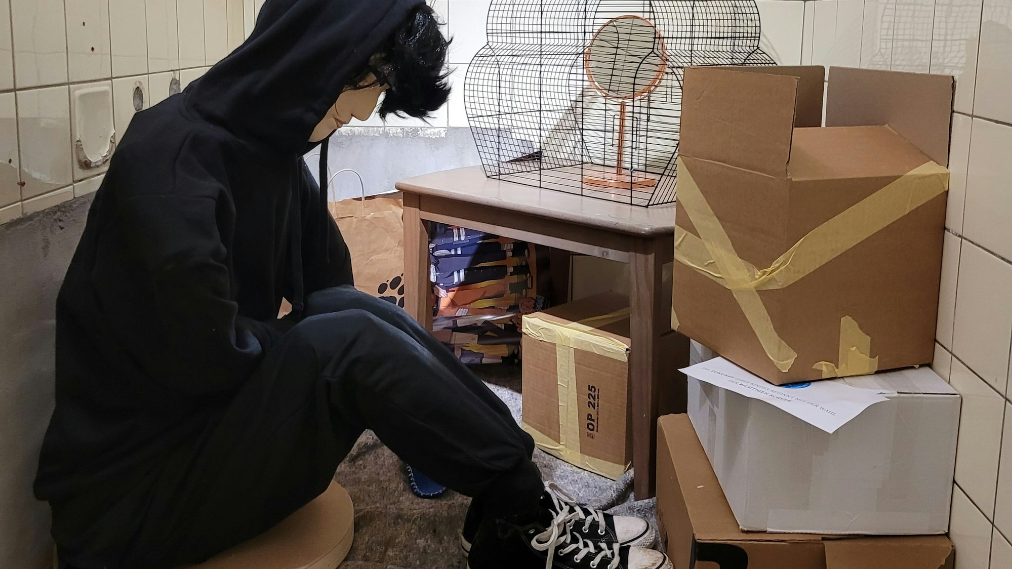 Installation: Im Vordergrund ein Jugendlicher, der auf dem Boden sitz, umgeben von Kartons, einem Vogelkäfig. Er lässt den Kopf hängen.