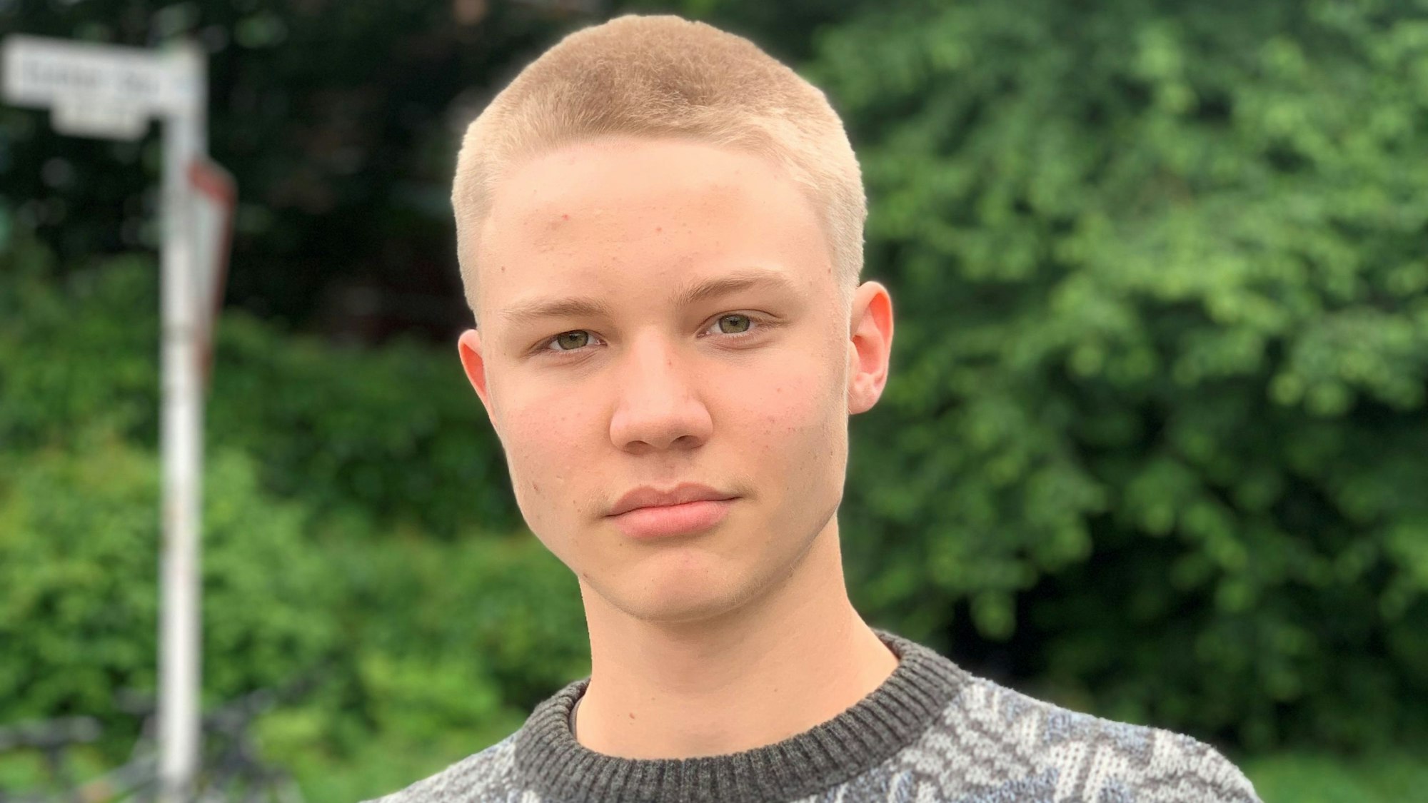 Ein junger Mann steht vor einer Grünanalge und lächelt.