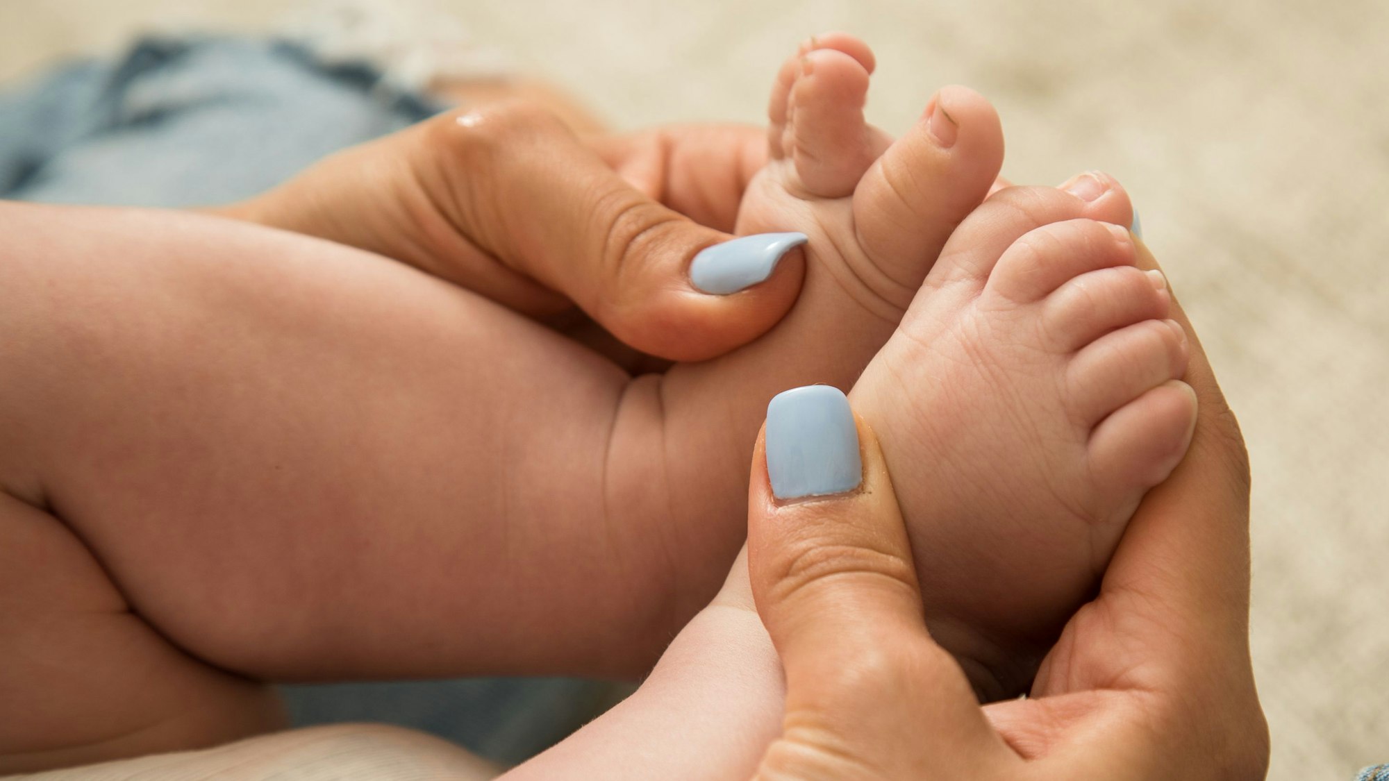 Eine Frau mit hellblau lackierten Fingernägeln hält die kleinen Füße eines Babys in ihren Händen.