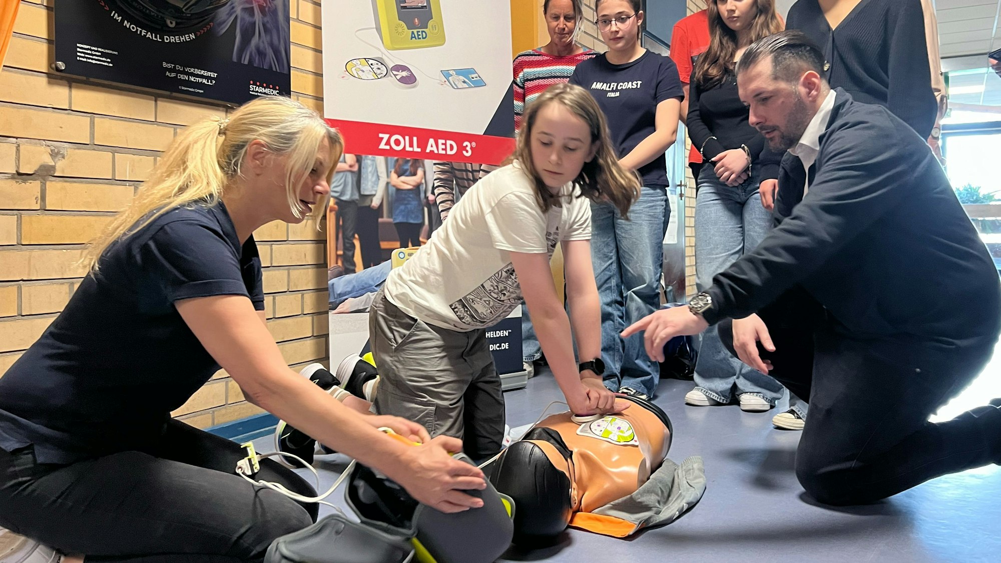 Matti (M.) probiert einen Defibrillator an einer Übungspuppe im Maximilian-Kolbe-Gymnasium in Porz-Wahn aus. Silke Voll (l.) von der Firma Zoll und Sascha Feldt (r.) von der Firma Starmedic geben Erklärungen. Schülerinnen und Lehrerinnen schauen zu.