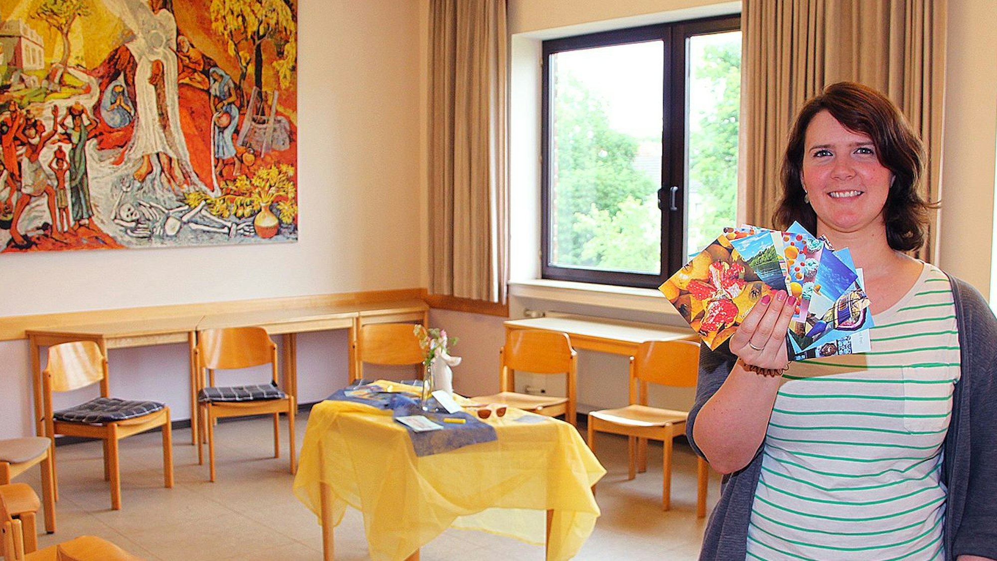 Linda Schmitt-Thees ist Pastoralreferentin in Mechernich. Sie ist rechts im Bild zu sehen und hält bunte Karten in der Hand. Daneben sind in einem Raum Stühle im Kreis um einen kleinen Tisch positioniert.
