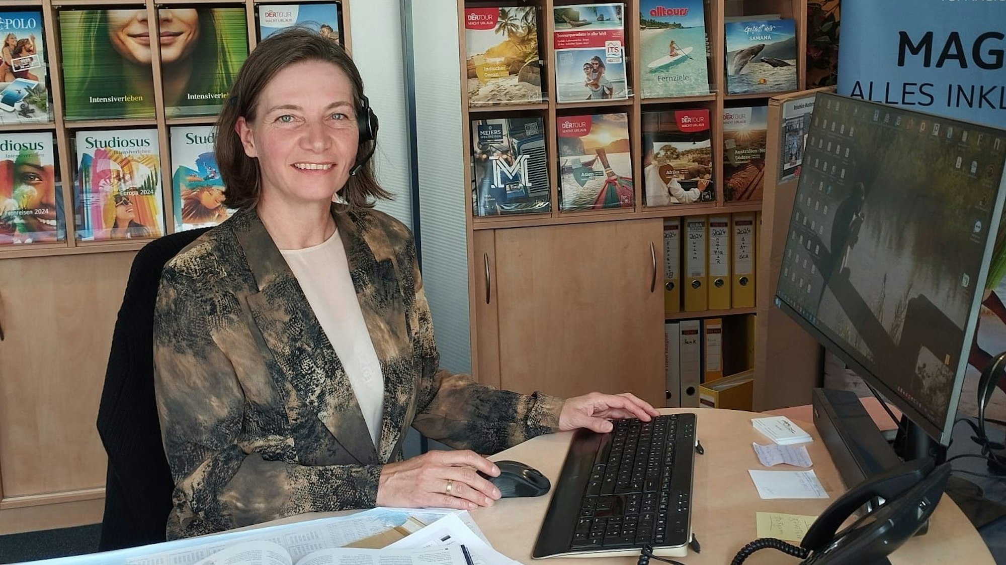 Eine Frau sitzt am Schreibtisch in einem Reisebüro. Im Regal stehen Kataloge.