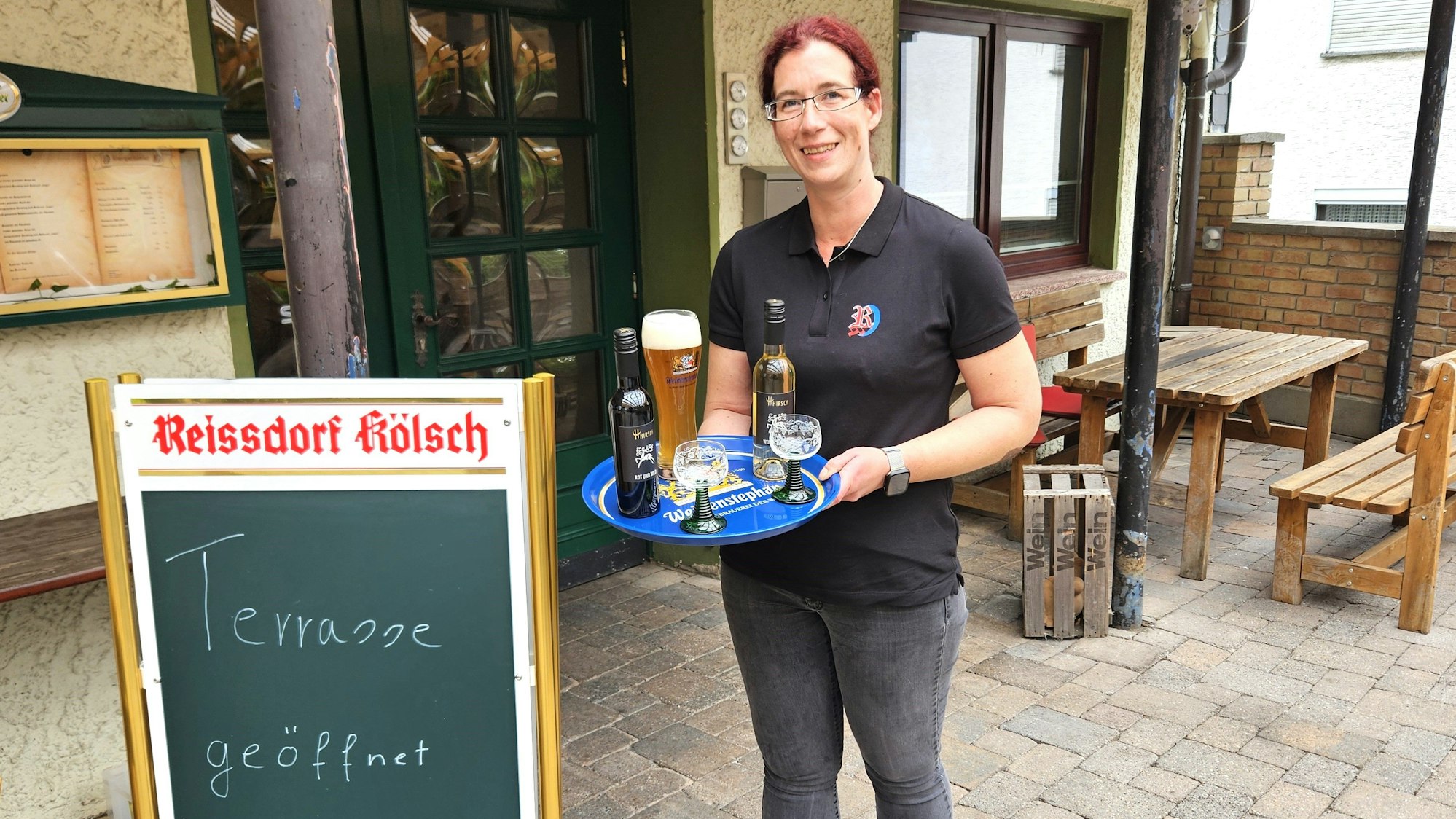 Regina Krüger trägt ein Tablett, auf dem ein Glas Weizenbier, eine Flasche Rotwein, eine Flasche Weißwein und zwei Weingläser stehen.