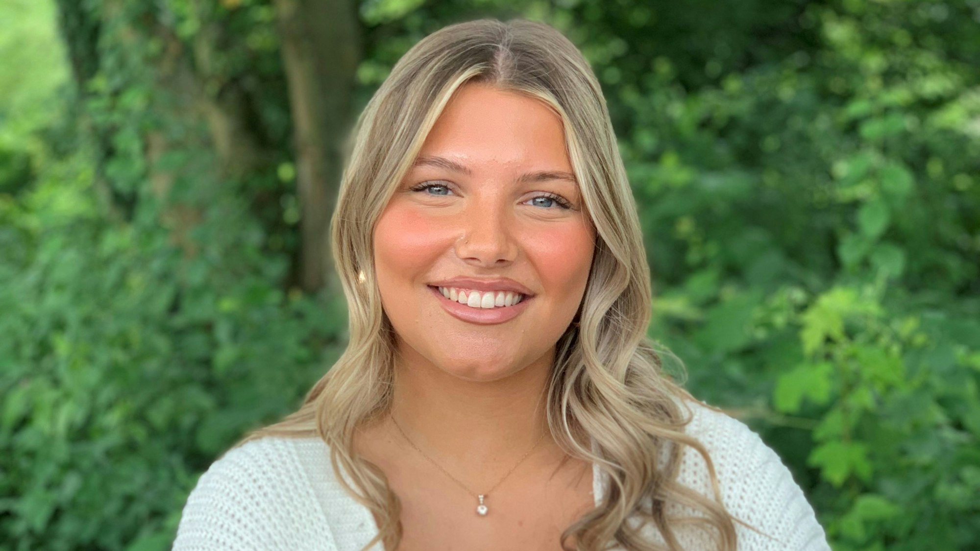 Eine junge Frau steht vor einer Grünanlage und lächelt.