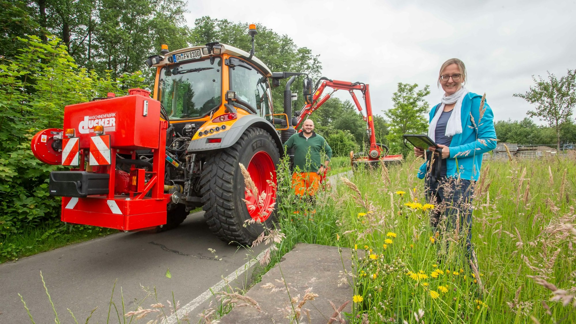 Manuela Thomas steht mit einem Tablet an einem dicht bewachsenen Wegesrand. Hinter ihr steht Christoph Seelbach neben einer großen Nähmaschine.