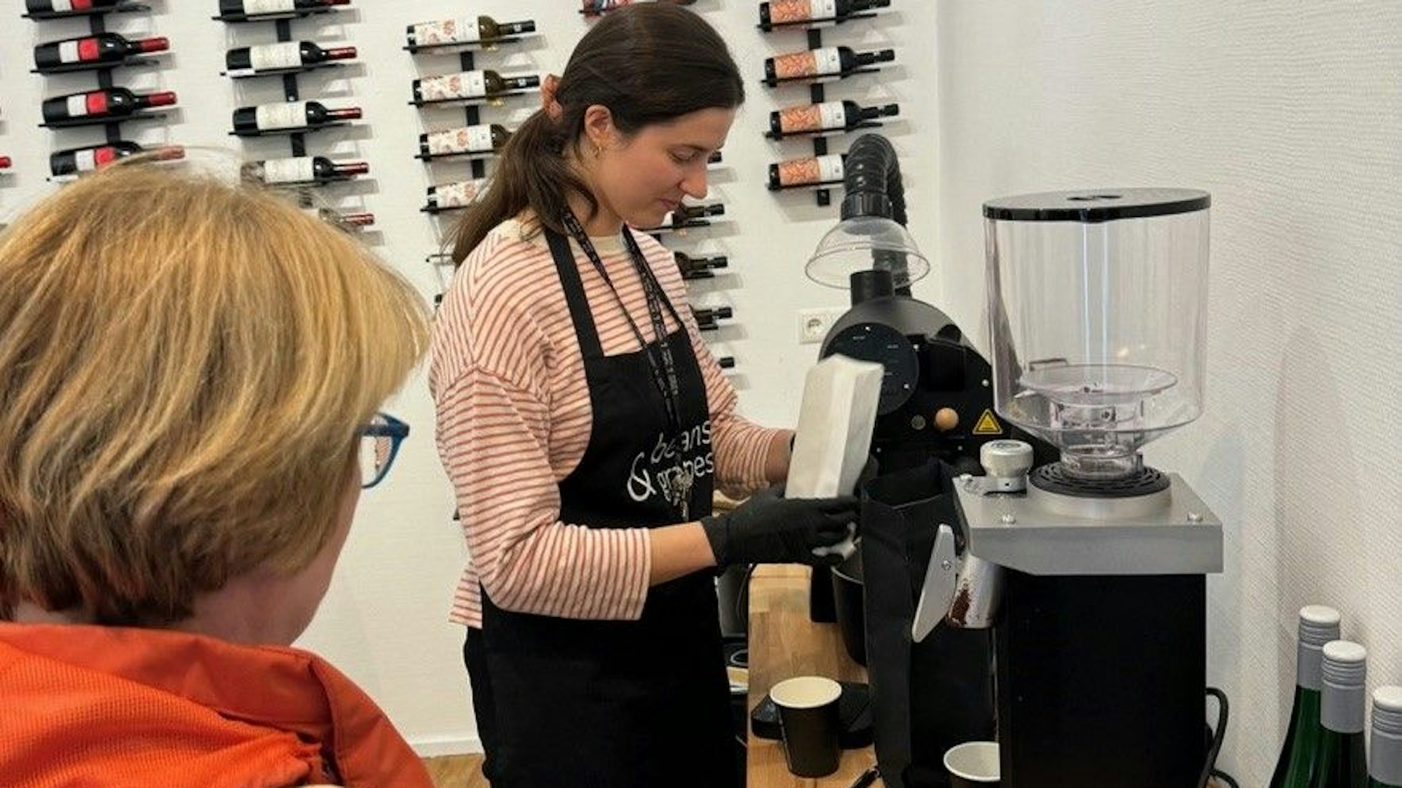 Eine Person steht an einer Kaffeemühle. Im Vordergrund eine weitere Person. Im Hintergrund sind Weinflaschen an der Wand aufgereiht.