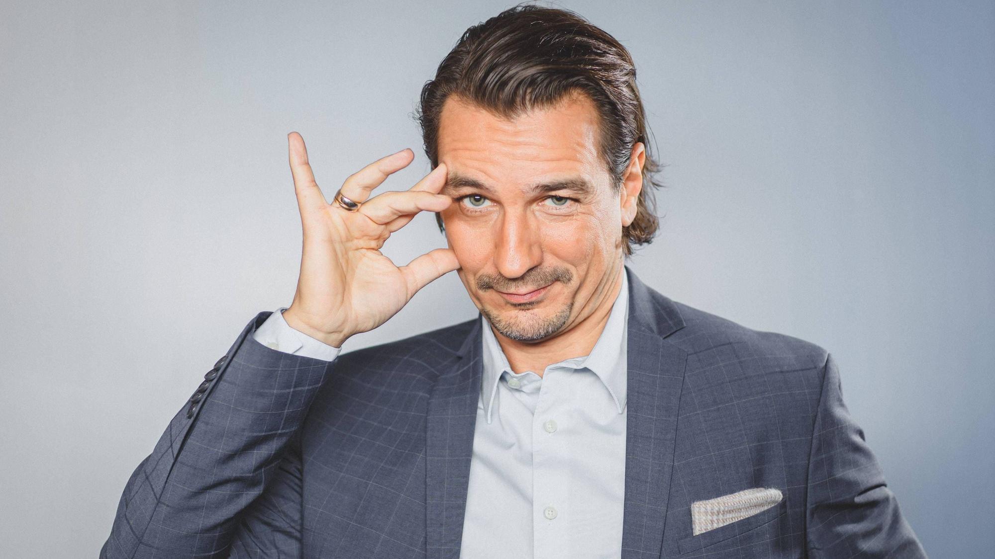 Mann mit Anzug, Hemd, und zurückgegeltem braunen Haar, der sich ans mit der Hand ans Gesicht fasst und leicht schmunzelnd
