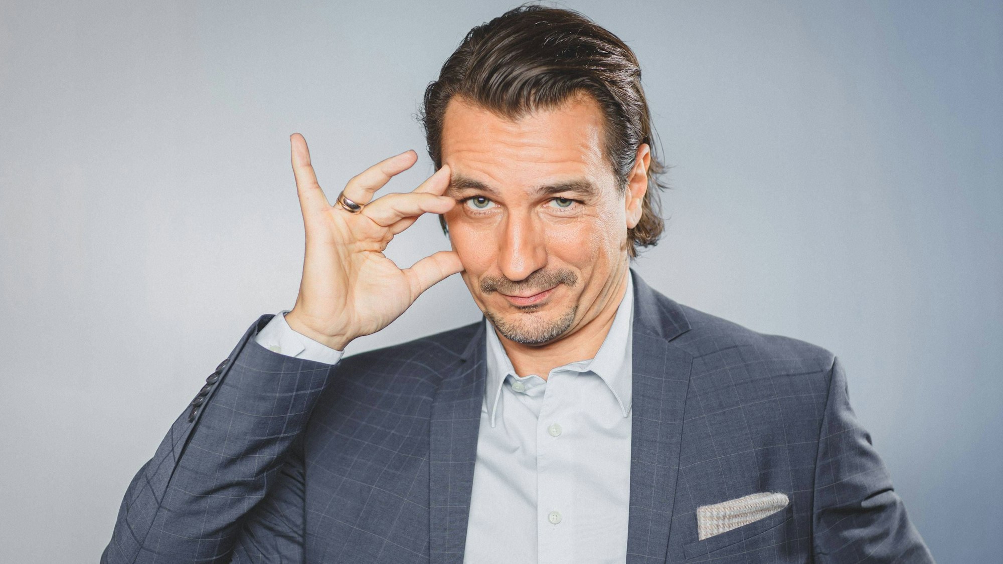 Mann mit Anzug, Hemd, und zurückgegeltem braunen Haar, der sich ans mit der Hand ans Gesicht fasst und leicht schmunzelnd