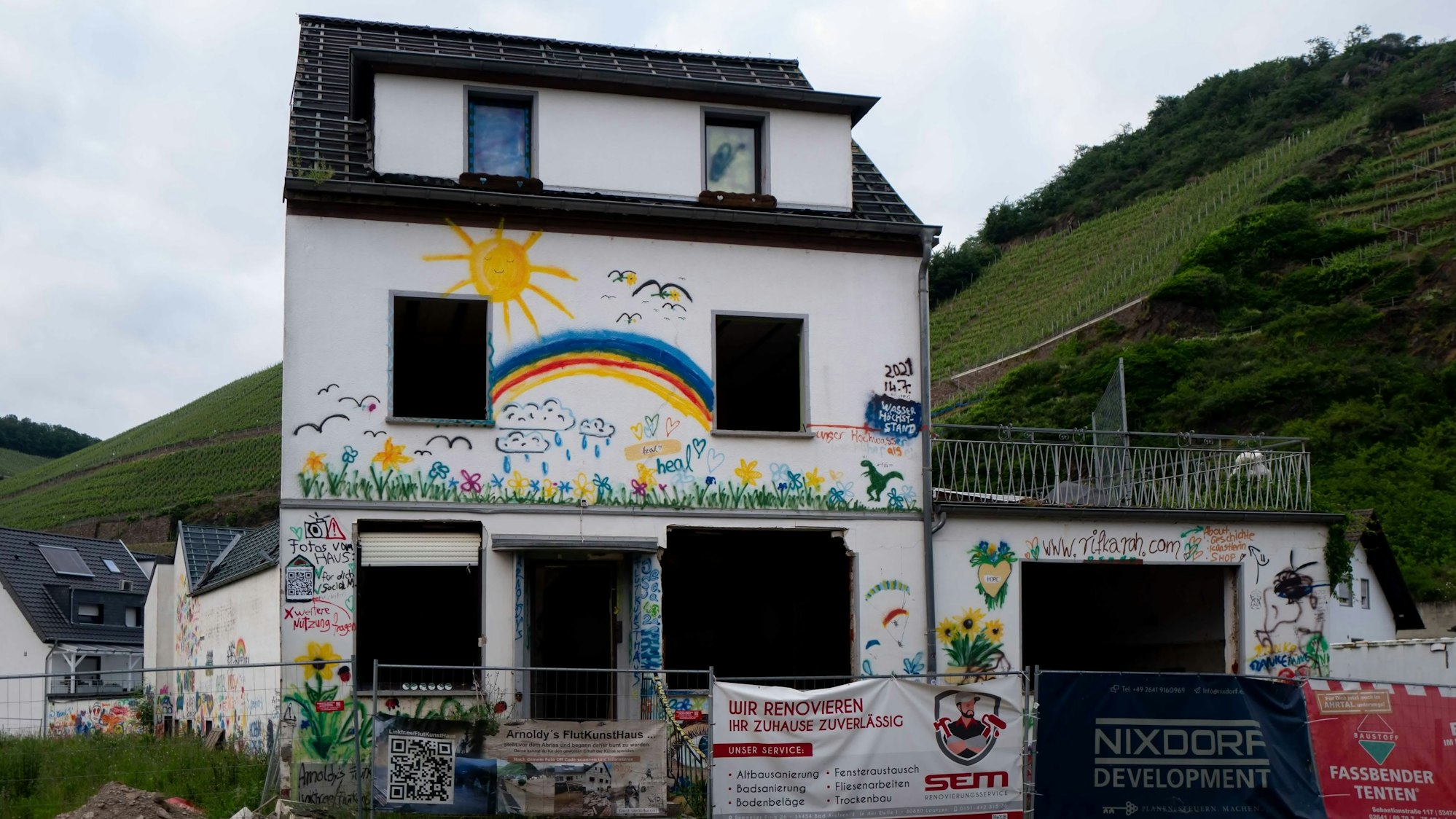 Eines der Häuser in Dernau an der Ahr, die bei der Flut 2021 zerstört wurden, ist zum Symbol der Katastrophe geworden. Auf die weiße Fassade haben Helfer unter anderem bunte Blumen, einen Regenbogen und eine gelbe Sonne gemalt.