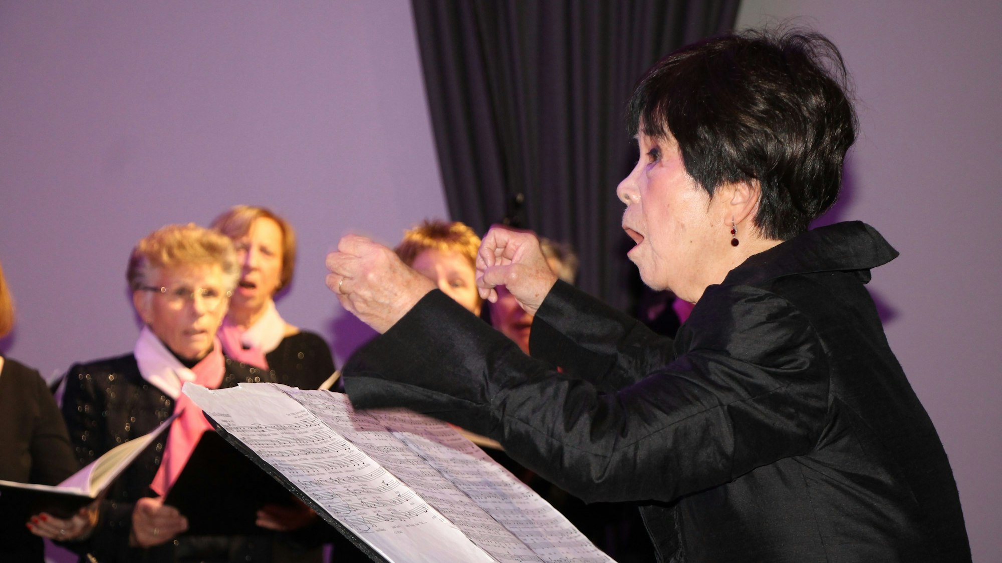 Eine Frau vor einem Notenblatt dirigiert mit ausladender Geste einen Frauenchor.